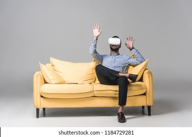 bearded man gesturing and using virtual reality headset and sitting on yellow sofa on grey