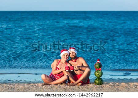 bearded man and girl in Santa clothes are sitting on sea beach with cups in their hands and snowman from watermelons. Summer Christmas Celebration