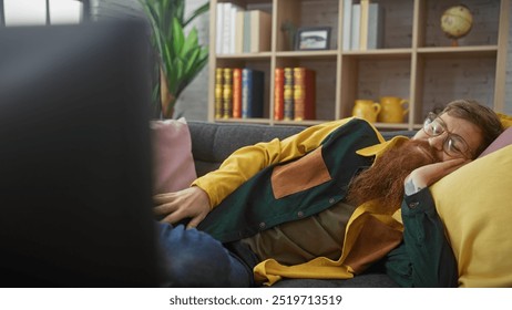 A bearded man with glasses and a casual outfit lounges on a sofa indoors, looking relaxed with a contemplative expression. - Powered by Shutterstock