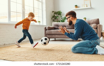 Bearded Man In Glasses Kneeling On Floor And Catching Ball While Teaching Boy To Play Football In Cozy Room At Home
