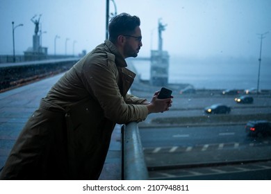 A Bearded Man With Glasses In A Winter Coat, Standing Leaning On The Railing Of A Bridge Over A Canal, Deep In Thought, Looking Straight Ahead.