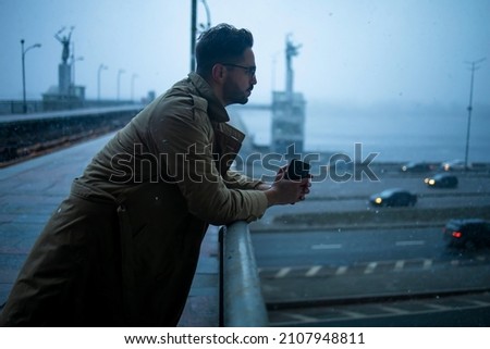 A bearded man with glasses in a winter coat, standing leaning on the railing of a bridge over a canal, deep in thought, looking straight ahead.