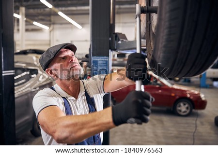 Bearded man in gloves tightening bolts on wheel with torque wrench while working in automobile repair garage