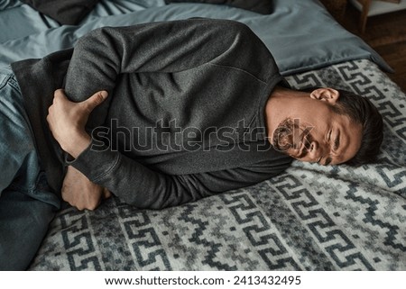 bearded man grimacing while lying on bed and suffering from stomachache, abdominal pain