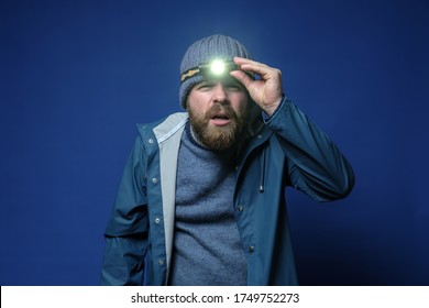 Bearded Man With A Headlamp In A Knitted Hat And A Waterproof Jacket Is Trying To See Something In The Light Of A Flashlight.