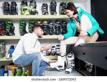 Bearded man helping smiling girlfriend to trying on ski boots in store of sports equipment