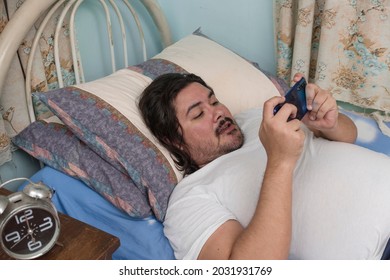 A bearded man in his 30s casually playing a MOBA game on his phone while lying in bed. Laziness or recreation during the weekend.
