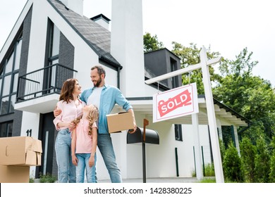 bearded man holding box and standing with wife and daughter near house and board with sold letters 