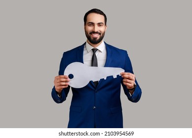 Bearded man holding in hand big paper key looking at camera with toothy smile, real estate, own accommodation, wearing official style suit. Indoor studio shot isolated on gray background. - Powered by Shutterstock