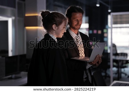 The bearded man holds the notebook while the young woman reads beside him, amber light strips warming the shadowy open plan interior in a bright modern coworking office.