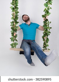 Bearded Man In Jeans, Swinging On Swings And Hamming To The Camera. Wooden Swing Suspended From A Rope Hemp, Rope Wrapped Vine And Ivy.