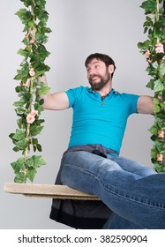 Bearded Man In Jeans, Swinging On Swings And Hamming To The Camera. Wooden Swing Suspended From A Rope Hemp, Rope Wrapped Vine And Ivy.