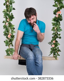 Bearded Man In Jeans, Swinging On Swings And Hamming To The Camera. Wooden Swing Suspended From A Rope Hemp, Rope Wrapped Vine And Ivy. Different Emotions