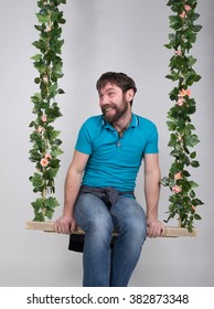 Bearded Man In Jeans, Swinging On Swings And Hamming To The Camera. Wooden Swing Suspended From A Rope Hemp, Rope Wrapped Vine And Ivy. Different Emotions