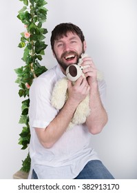 Bearded Man In Jeans And A White Shirt, Hugging A Teddy Bear, Swinging On Swings And Hamming To The Camera. Wooden Swing Suspended From A Rope Hemp, Rope Wrapped Vine And Ivy