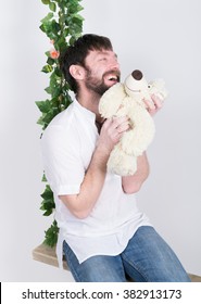 Bearded Man In Jeans And A White Shirt, Hugging A Teddy Bear, Swinging On Swings And Hamming To The Camera. Wooden Swing Suspended From A Rope Hemp, Rope Wrapped Vine And Ivy