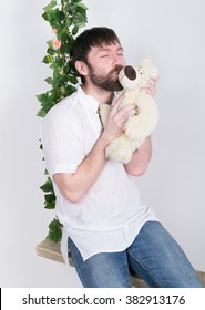 Bearded Man In Jeans And A White Shirt, Hugging A Teddy Bear, Swinging On Swings And Hamming To The Camera. Wooden Swing Suspended From A Rope Hemp, Rope Wrapped Vine And Ivy