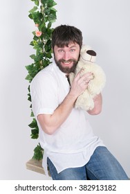 Bearded Man In Jeans And A White Shirt, Hugging A Teddy Bear, Swinging On Swings And Hamming To The Camera. Wooden Swing Suspended From A Rope Hemp, Rope Wrapped Vine And Ivy