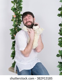 Bearded Man In Jeans And A White Shirt, Hugging A Teddy Bear, Swinging On Swings And Hamming To The Camera. Wooden Swing Suspended From A Rope Hemp, Rope Wrapped Vine And Ivy