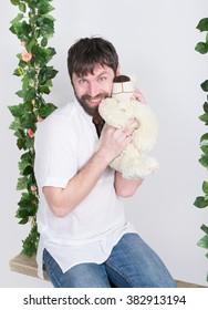 Bearded Man In Jeans And A White Shirt, Hugging A Teddy Bear, Swinging On Swings And Hamming To The Camera. Wooden Swing Suspended From A Rope Hemp, Rope Wrapped Vine And Ivy