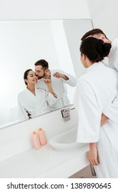 Bearded Man Kissing Cheek Of Cheerful Woman Looking At Mirror In Bathroom 