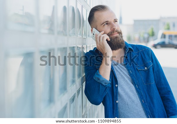 Bearded man leaning\
on a wall in a city street chatting on a mobile phone looking up as\
he listens to the call