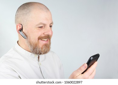 Bearded Man In A Light Shirt With A Mobile Phone And Bluetooth In His Ear
