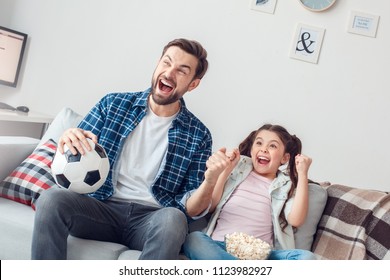 Bearded man and little girl at home family time sitting on sofa watching football hands up screaming cheerful goal