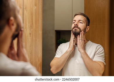 A bearded man is looking himself in the mirror and touching his beard.