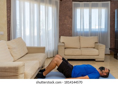 Bearded man in low shape exercising with black and blue sportswear and a mask to prevent coronavirus in the living room at home on a mat - Shutterstock ID 1814144570