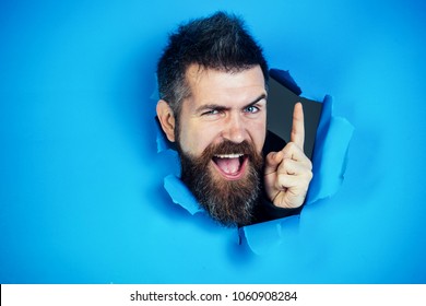 Bearded man making hole in paper. View of male face through hole in blue paper. Bearded man looking through hole and show the finger. Bearded man making hole in paper and points up.