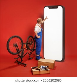 Bearded man, mechanic in blue overalls standing next to bicycle and interacting with giant mobile phone with white empty screen on red background. Concept of profession, occupation, repairing service - Powered by Shutterstock