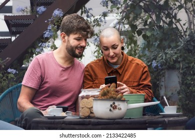 A bearded man and a nonbinary woman watching content online sitting on a terrace drinking coffee together - Powered by Shutterstock