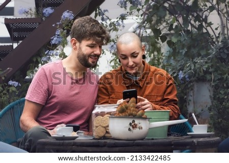 A bearded man and a nonbinary woman watching content online sitting on a terrace drinking coffee together