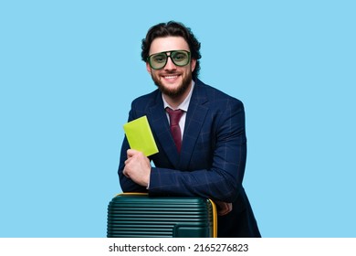 Bearded Man in Official Suit and Green Sunwear Traveling on Vacation Trip. Air Flight Business Class Journey. Happy Male Traveler Tourist holds Passport and Leans on Suitcase on Blue Background.