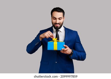 Bearded Man Opening Gift Box, Unwrapping Birthday Present For Celebrating Holiday, Pulling Ribbon, Looking At Gift, Wearing Official Style Suit. Indoor Studio Shot Isolated On Gray Background.