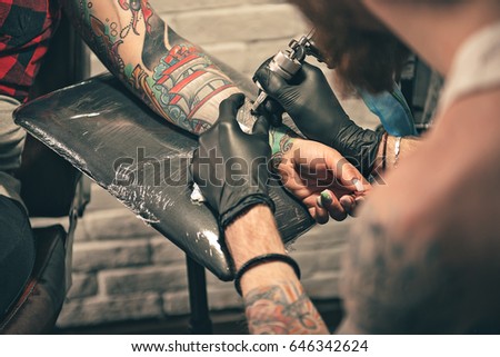 Bearded man painting image on arm of female with special equipment. He turning back to camera. Close up