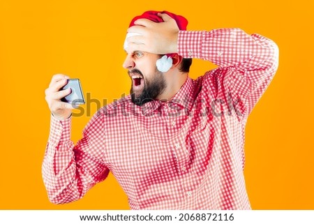 bearded man in a plaid shirt and Santa Claus hat is shocked and looks at the phone with surprise