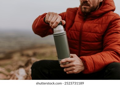 A bearded man in a red jacket opens a tumbler while sitting outdoors. Man with tumbler on outdoor adventure, hiking on mountain trail. Hiking on nature trail, outdoor activity. Adventure in nature.
