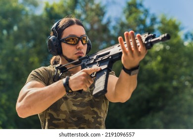 Bearded Man In Safety Goggles Headphones And Camo T-shirt Practicing Using Submachine Gun. Outdoor Firing Range. Horizontal Shot. High Quality Photo