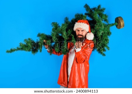Bearded man in Santa hat with Christmas tree sending air kiss. Christmastime. Delivery Santa man carrying pine tree on shoulder. Businessman with fir-tree blowing kiss. Winter holidays celebration.