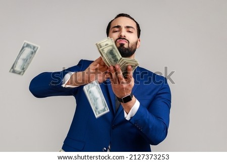 Bearded man scattering dollars with arrogant grimace, boasting wealthy life, concept of careless money spending, wearing official style suit. Indoor studio shot isolated on gray background.