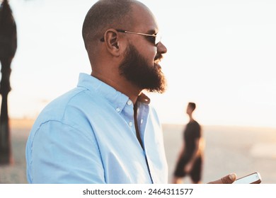 Bearded man sending text message via mobile telephone chatting online while enjoying summer evening, hipster guy satisfied with 4G connection in roaming share photos from vacation in social networks