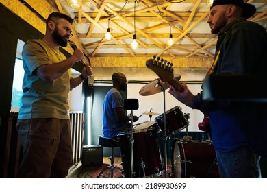 Bearded man singing in microphone among his buddies playing electric guitar and hitting drum set during repetition in garage - Powered by Shutterstock