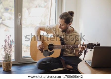 bearded man is sitting at home next to the window and playing the acoustic guitar