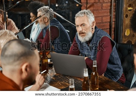 bearded man sitting near laptop and discussing startup project with multiethnic team in bar
