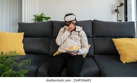 Bearded Man Sitting On Couch And Watching Movie With VR Glasses Headset While Eating Popcorn At Cozy Home. Enjoy Immersive Futuristic Virtual Reality 3D Travel Tour, Watch 360 Video