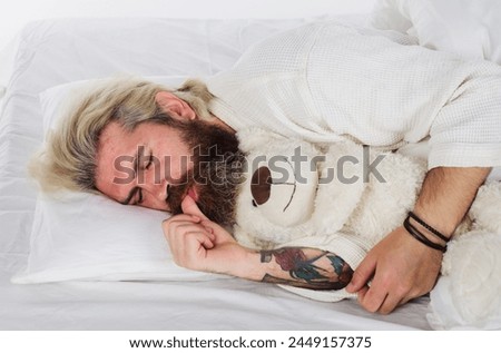 Bearded man sleeping with plush teddy bear relaxing in bed. Good night. Sleepy man in pajamas sleeping in bed at home with teddy bear toy. Handsome guy sleeping at bedroom and hugging soft teddybear.