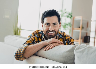 A bearded man smiles while leaning on a sofa in a sunlit living room, conveying warmth, comfort and relaxed home lifestyle perfect for lifestyle, wellness and interior concepts.