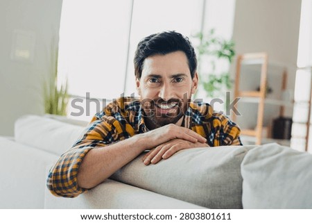 A bearded man smiles while leaning on a sofa in a sunlit living room, conveying warmth, comfort and relaxed home lifestyle perfect for lifestyle, wellness and interior concepts.